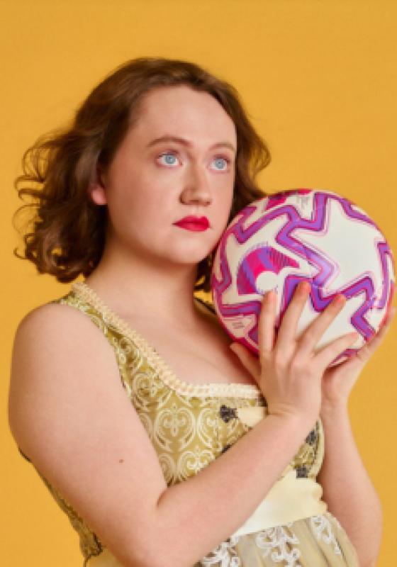 Dee stands in front of a yellow back drop. They are holding white, pink and purple football and gazing into the distance.