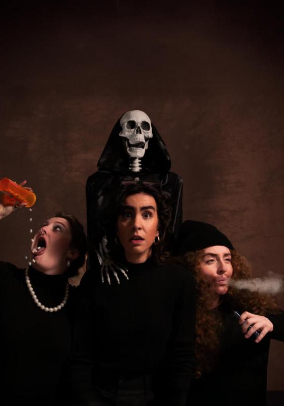 Three performers stand before a skeleton in a robe: the one in the center looks straight ahead, the one on their left smokes, and the one on their right pours a bottle of pills into their mouth, in front of a brown background.