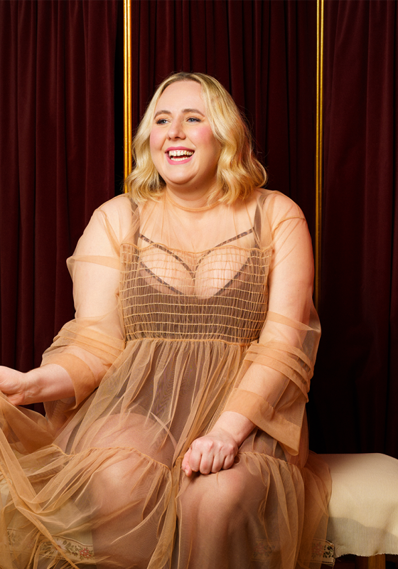Helen sits on a bench, dressed in a sheer flowy dress. They smile happily. 