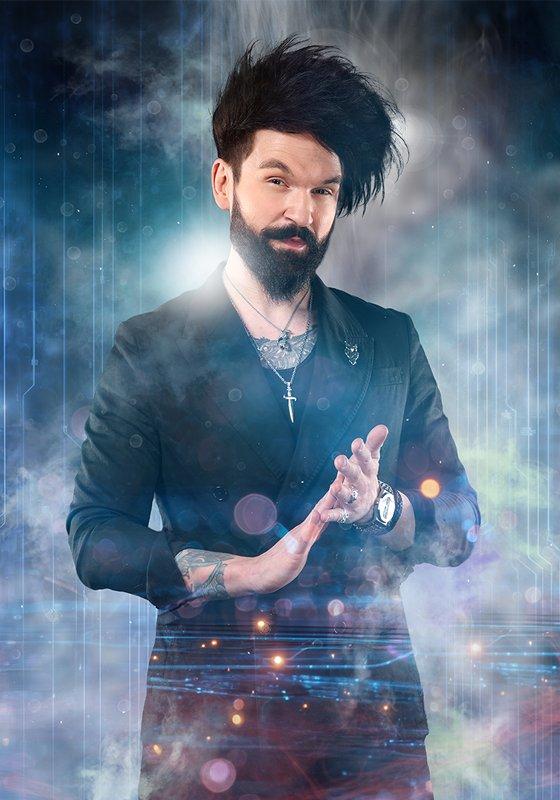 Colin Cloud, wearing a black suit jacket with chain necklaces, rubs his fingertips of one hand onto the palm of his other. A ball of light emanates from his palm. He is surrounded by smoke. 