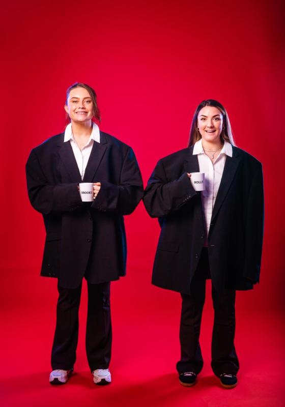 Holly and Brooke stand against a red background, wearing oversized suits