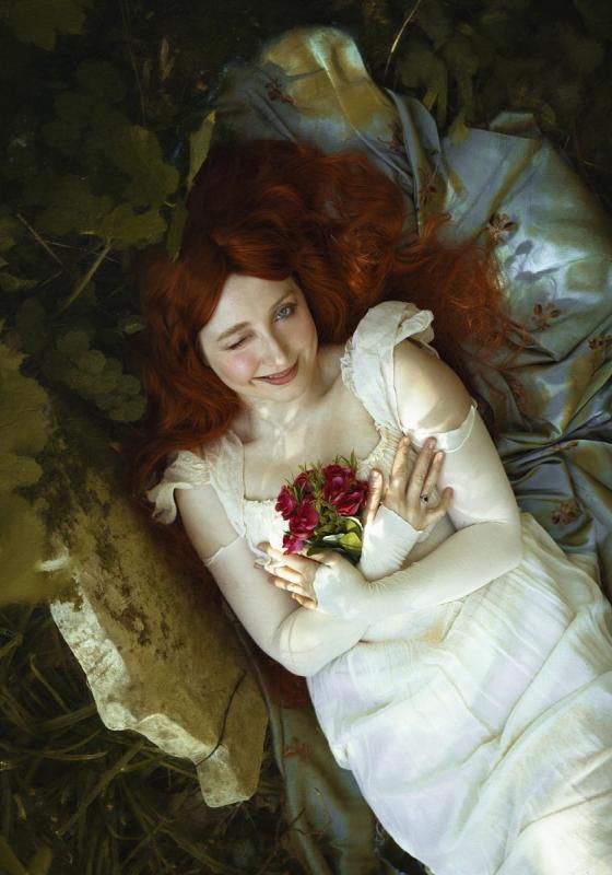 Bebe laying in what appears to be some kind of casket with her red hair flowing against the leaves and holding some red flowers. She may be laying in a grave.