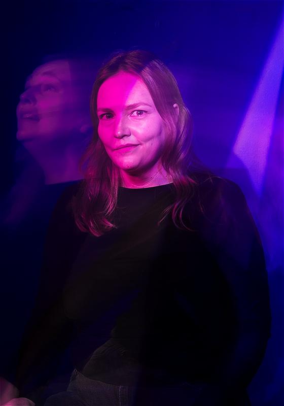 Kate is looking into the camera. A beam of light illuminates half of her face. 