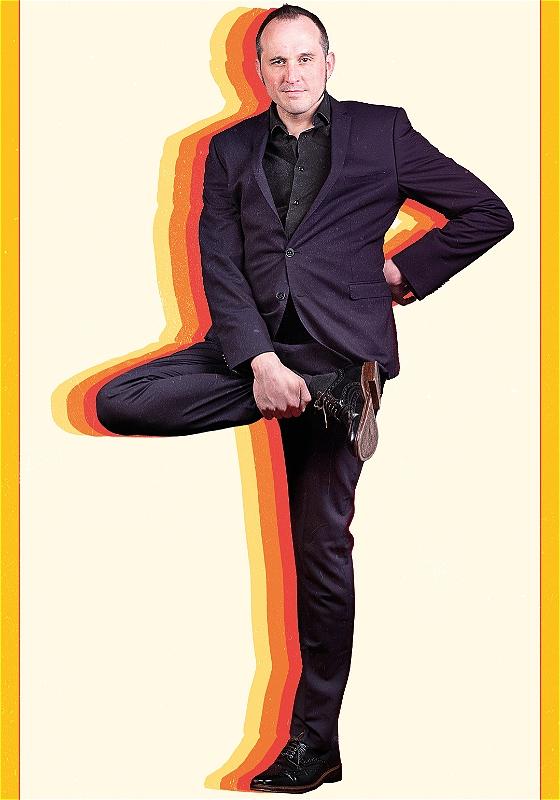 Phil looks into the camera whilst holding up their right foot. Behind them are colourful silhouettes. 