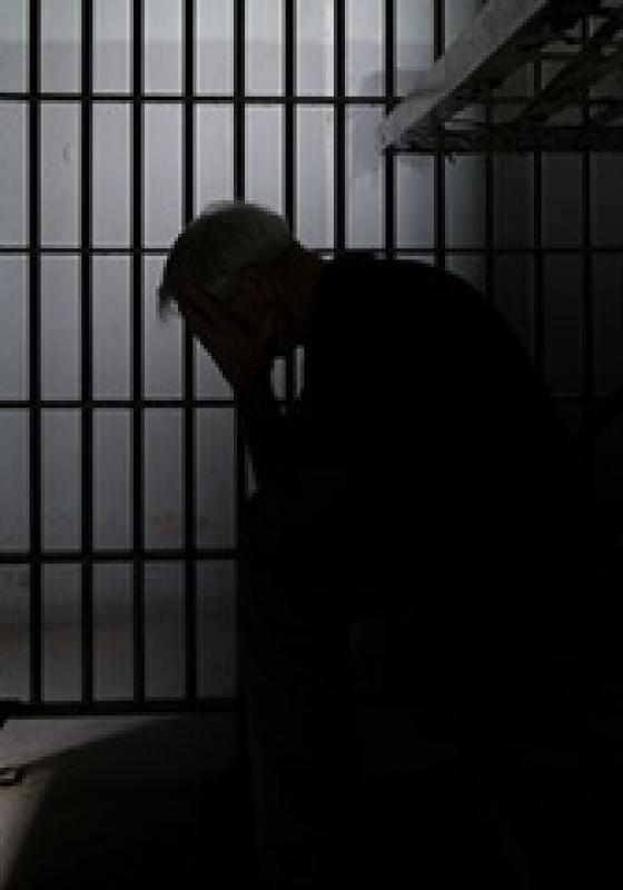 A person sits in a dark cell with their head in their hands