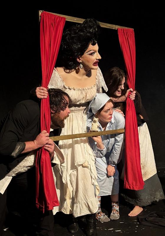 The performers huddle together around a window frame prop in front of a black background