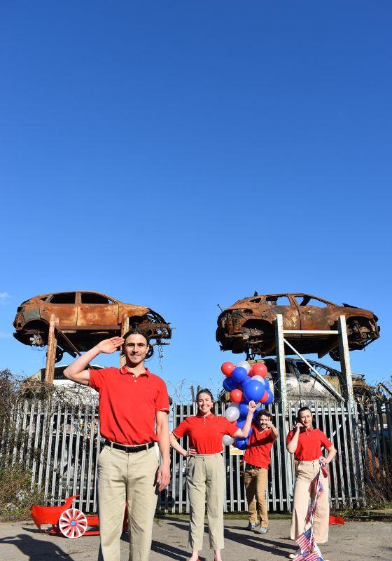 Four performers wearing red polos and khaki pants stand in front of a car junkyard