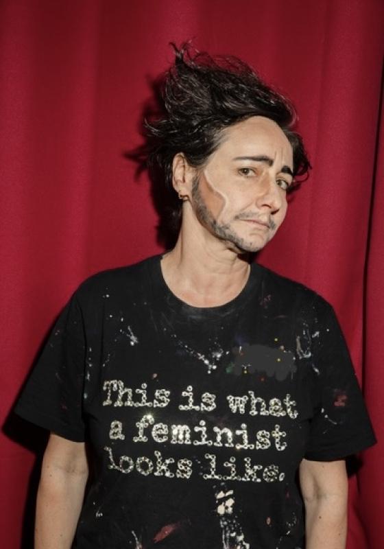 The performer stand before a red velvet curtain wearing a paint-splattered shirt that says "this is what a feminist looks like." in rhinestones.