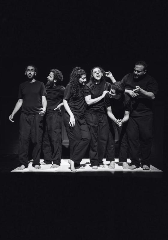 A group of performers wearing black stand on a white stage, surrounded by darkness. 