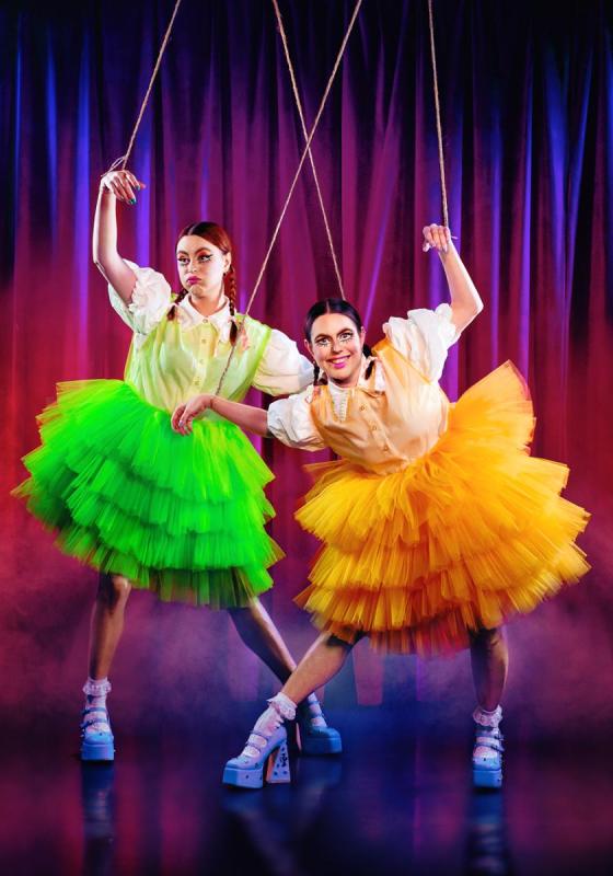 Maddy and Marina Bye are attached to strings, their arms being pulled up above their heads. They are wearing a bright green and orange tutu respectively