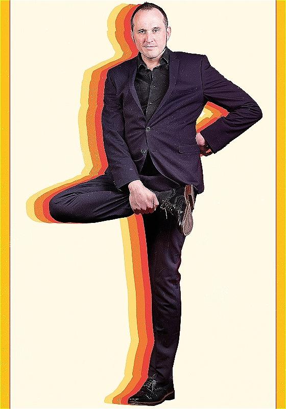 Phil looks into the camera whilst holding up their right foot. Behind them are colourful silhouettes. 