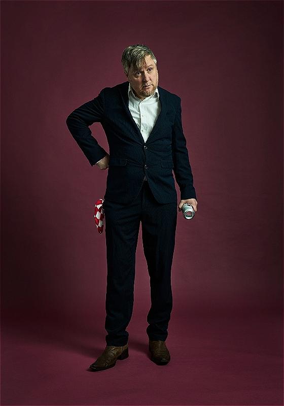 Tim Key stands in a suit with his hand on his hip against a purple backdrop