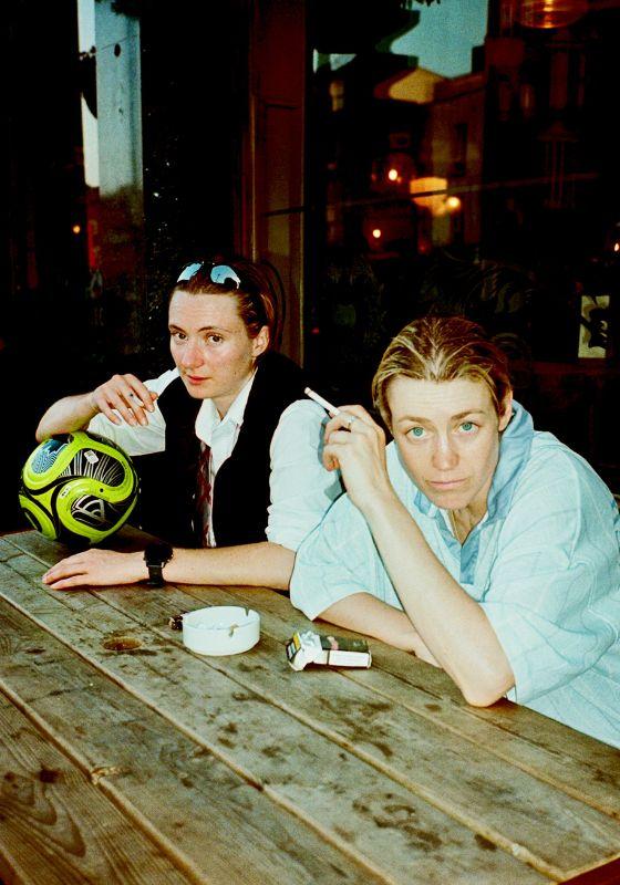 The performers sat outside a pub smoking and the one holding a football. The title is written above in lime green.