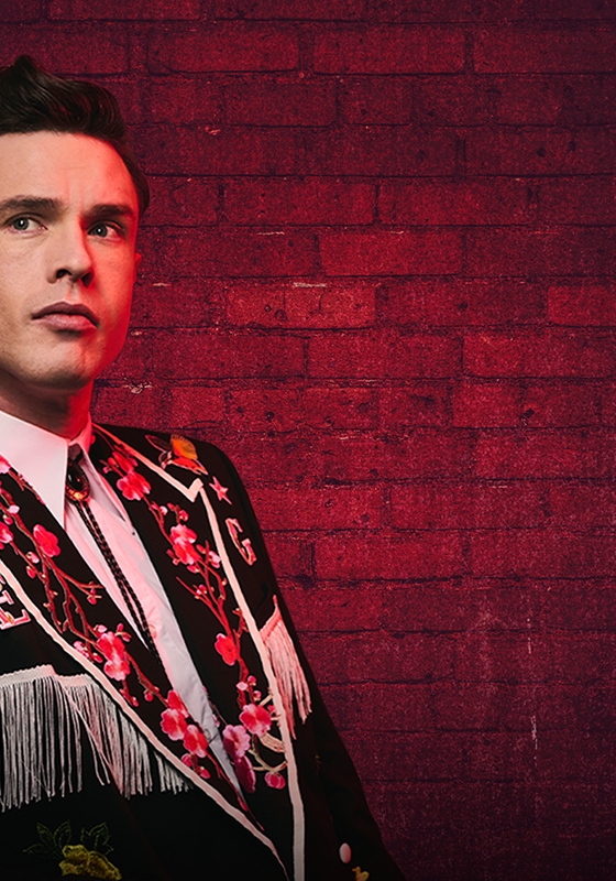 Ed Gamble stands in front of a red brick wall. He is dressed in a suit decorated with flowers and fringe.