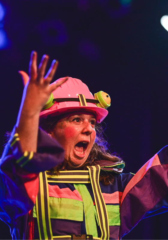 A woman is wearing a colourful costume and a hard hat that resembles a frog. She stands with her hands in the air with a shocked expression.