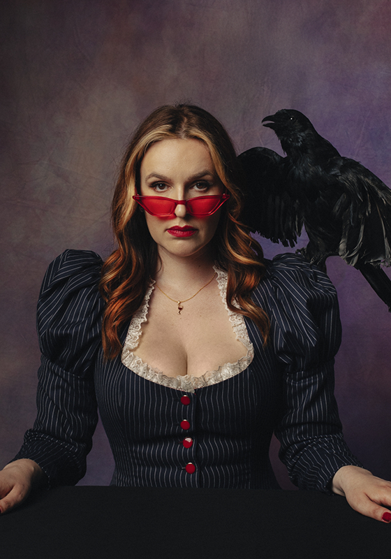 Katie Norris, a white woman with brown hair, stares into the camera. She is wearing a pair of red tinted sunglasses and a crow is perched on her shoulder.