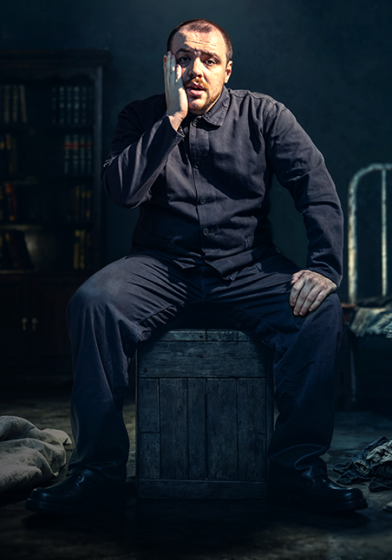 A person is in a dimly lit cell, sitting on a crate. They're wearing all-black and they look worried, holding a hand up to their bearded face. 