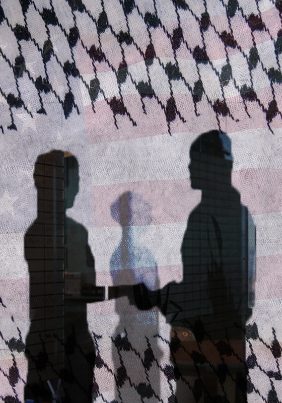 The shadows of two people holding hands. They can be seen in a cutout of fencing. In the background is a faded American flag.