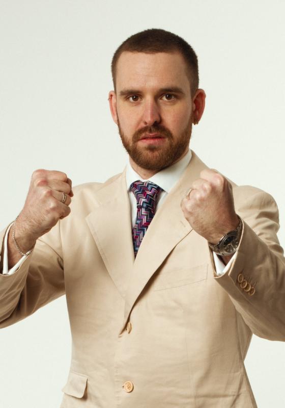 Rob, in a pink suit jacket against a white background, holding up his fists combatively 