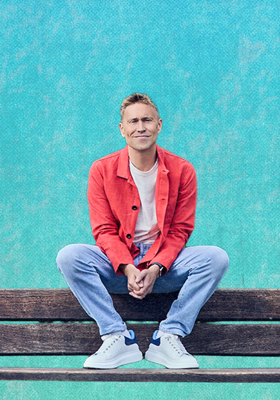 Russell Howard sits on the back of a bench in front of a blue backdrop. He has an amused expression.