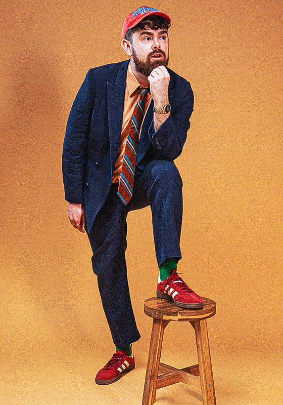 Tom is dressed in a navy suit, orange shirt, striped tie and red cap. He stands against an orange background with his foot perched on a wooden stool.