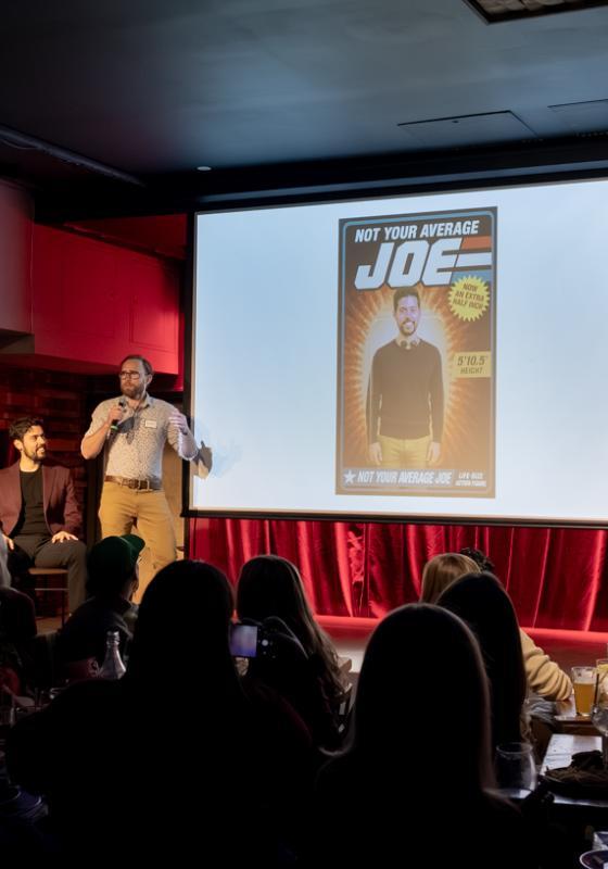 Three men standing on a stage. One of the men is holding a microphone and pitching his friend to the audience using a powerpoint presentation.