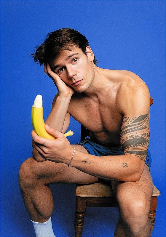 Mario Adrion sits shirtless in front of a blue background holding a partly peeled banana 