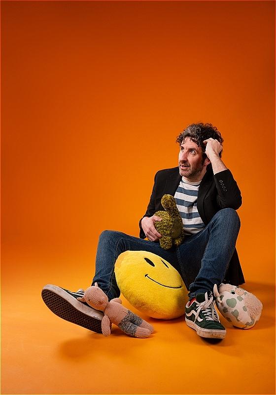 Mark Watson sitting on the floor holding children's toys and stuffed plushies.