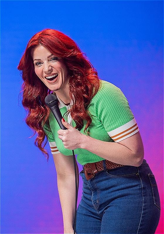 A woman dressed in a green shirt and blue jeans laughs into a microphone. Behind her is a bright pink and blue backdrop.