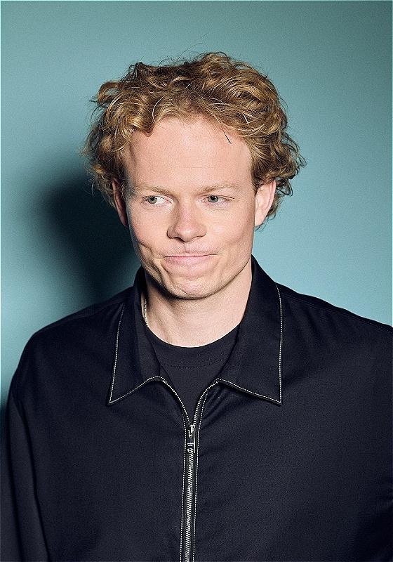 A curly haired man wearing a black zip up jacket gazes down with a mildly thoughtful expression. He is stood in front of a blue wall. 