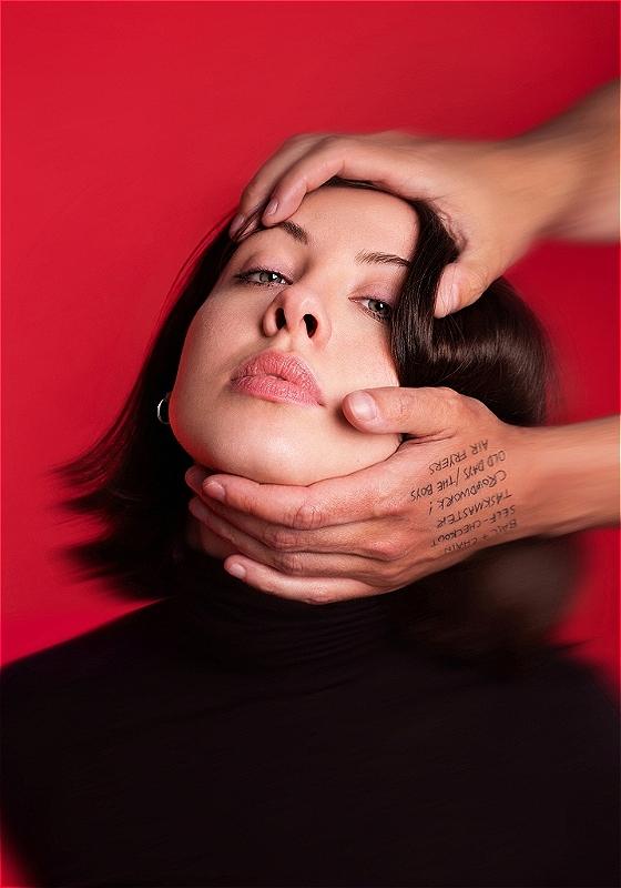A blurry image of Harriet's face against a red background. Someone's hands reach in from out of frame and are holding the top of her head and her chin, pulling her head back so her face is tilted upwards