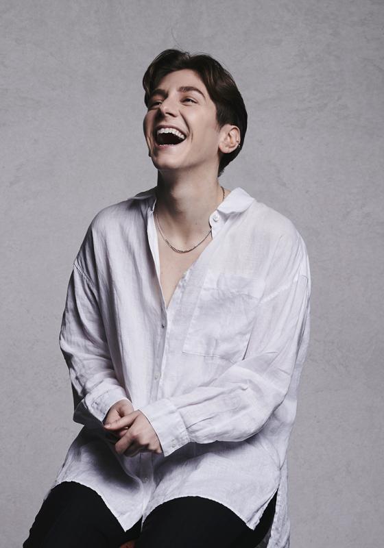 Sarah laughing on a stool wearing an oversized white shirt. There is a grey background.