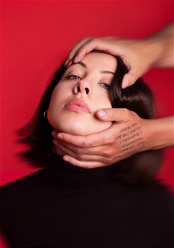 A blurry image of Harriet's face against a red background. Someone's hands reach in from out of frame and are holding the top of her head and her chin, pulling her head back so her face is tilted upwards