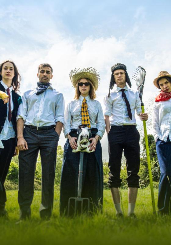 5 young adults in professional attire stand in a field holding farming equipment and looking at the camera.
