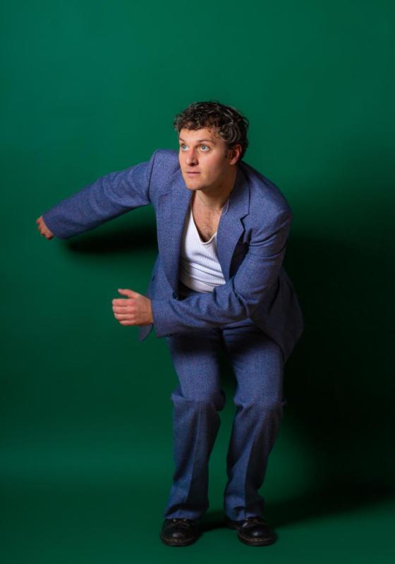 Jonathan wearing a blue blazer and trousers over a white t shirt crouches in front of a green backdrop. He is swinging his arms to one side and looking off camera to the left. 