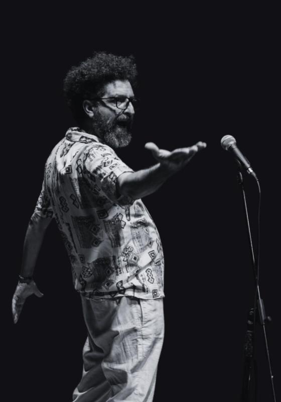 A black and white image of the performer standing in front of a microphone. They are reaching out their hand in a powerful pose.