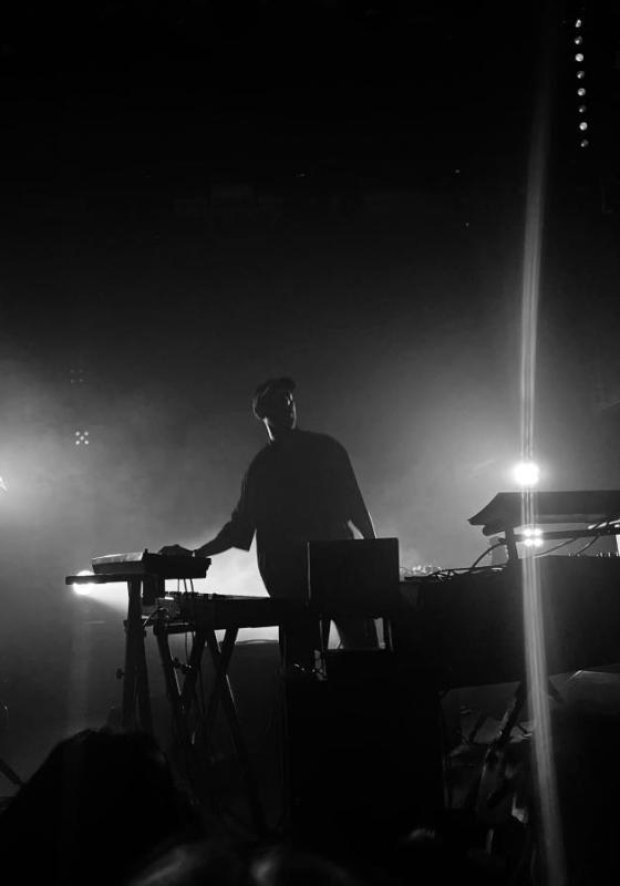 Isam stands in front of keyboards and other musical instruments, on a dark stage with bright stage lights