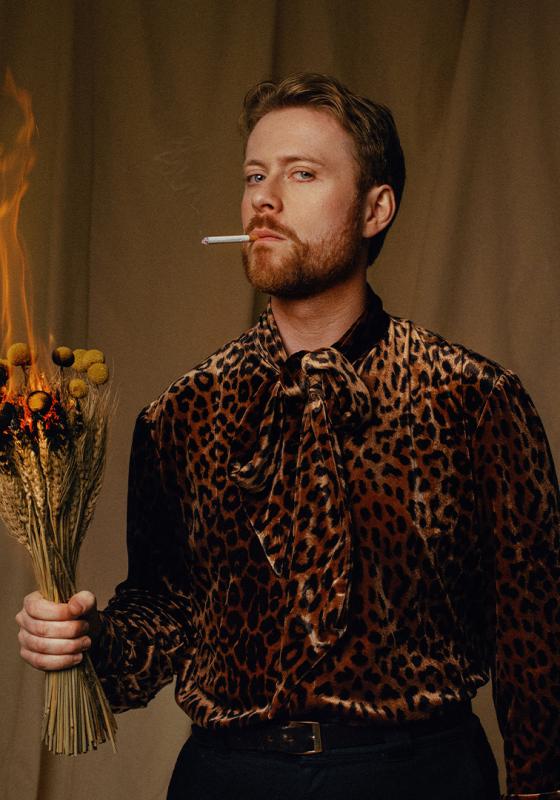 John wears a leopard print shirt and looks at the camera with a cigarette in their mouth. They are holding a bouquet of flowers that is on fire. 