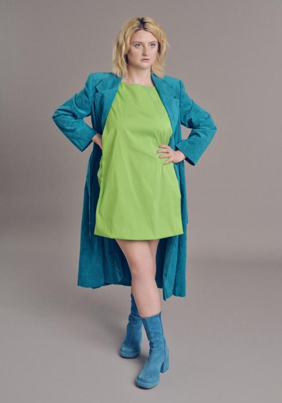 Lorna stands posing with their hands on their waist. They are wearing a green dress and a blue jacket and boots. 