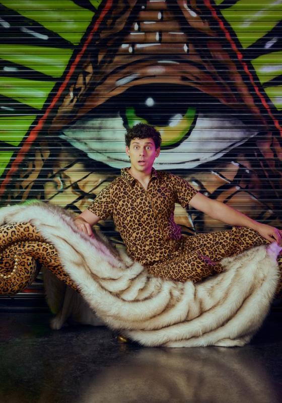 Matt wearing leopard print shirt and trousers, sat on a tiger print blanket on a leopard print chaise lounge. The chaise lounge is in front of a shutter graffiti-d with an eye in a triangle. 