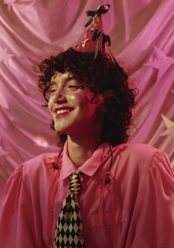 The performer, dressed in a pink shirt, tie and party hat, smiles joyfully in front of a pink back drop.