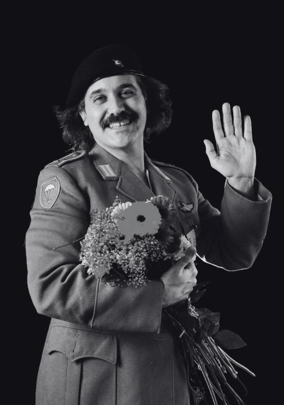 A black and white image of the performer wearing uniform and holding flowers waves at the camera with a large smile on their face