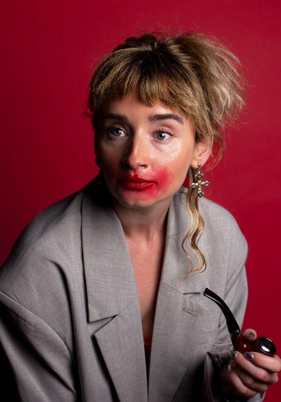 Molly is sitting against a red backdrop. She's wearing a grey blazer and holding a pipe. She has bright red lipstick smeared across her blank expression.