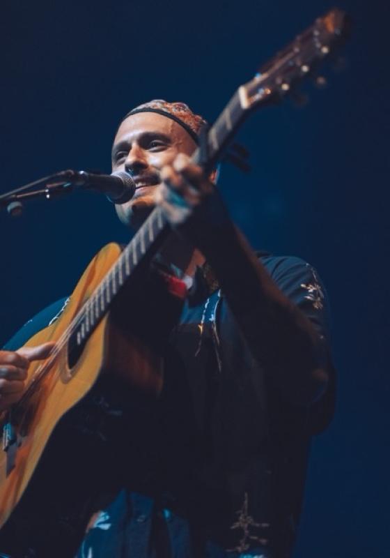 Ahmed stands behind a microphone on stage, playing a guitar