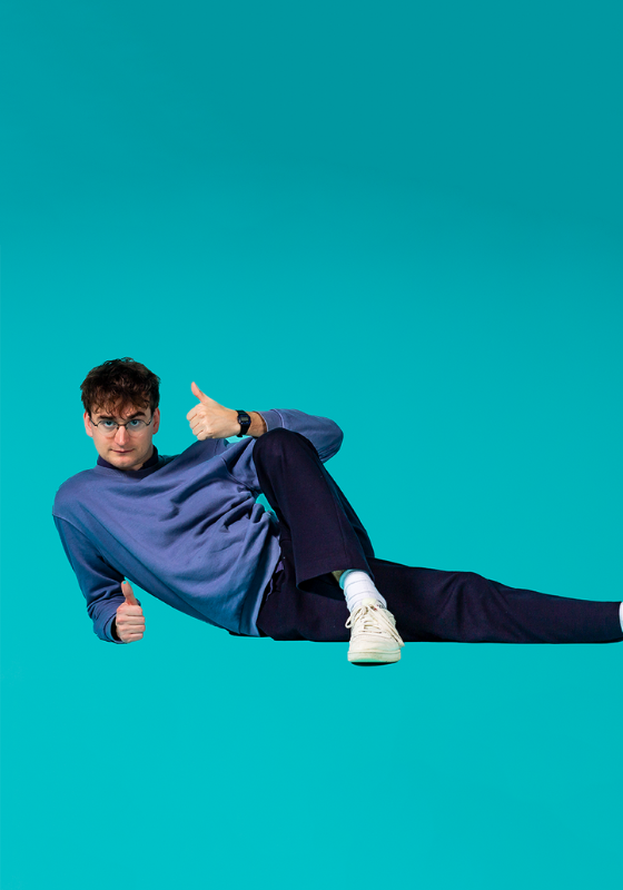 Sam lying on the floor holding a thumbs up against a bright blue background.