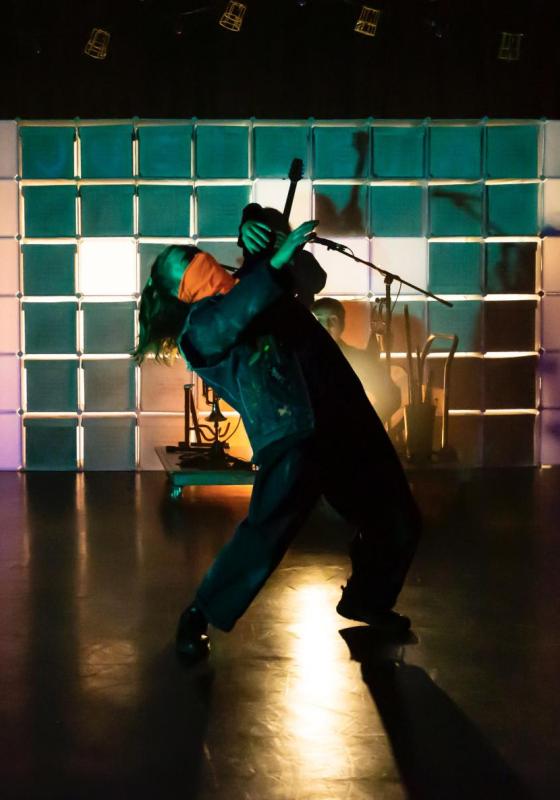 The performer, dressed all in green, leans back whilst stood under a spotlight on stage.