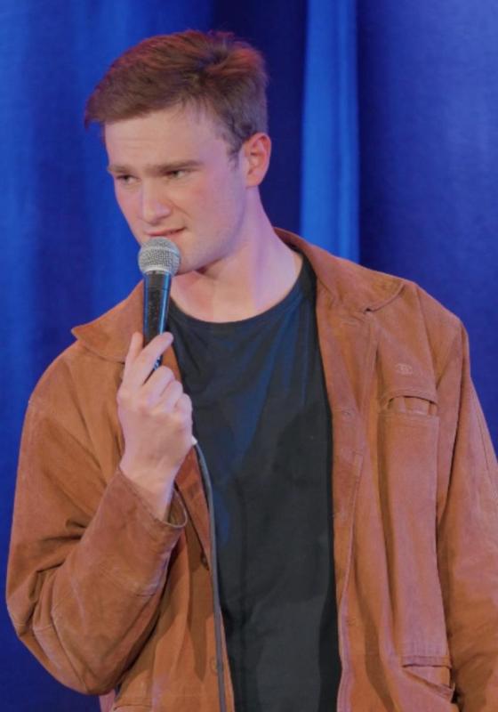 Josh Berry standing on stage holding a microphone against a blue curtain
