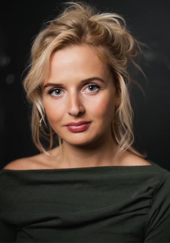 Headshot of Alana. Blonde hair, large gold hoops earrings and a slash neck grey top.