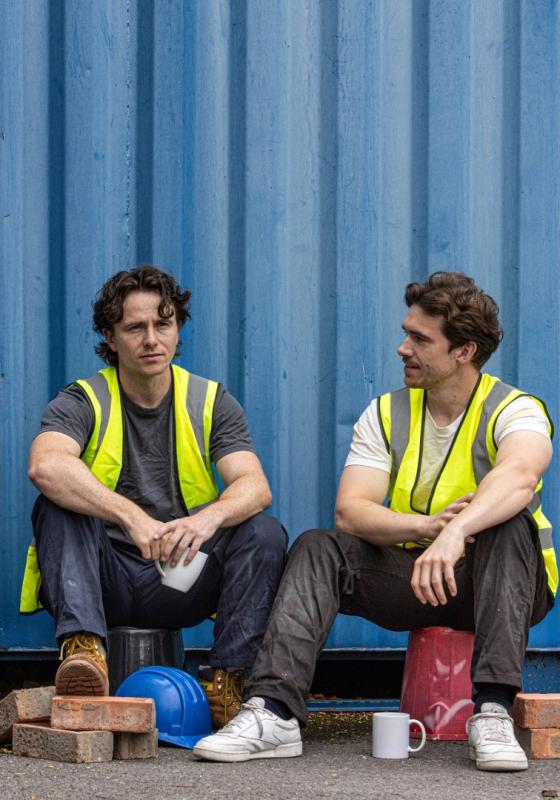Two people are sat on buckets. They are both wearing high vis vests and are surrounded by building bricks