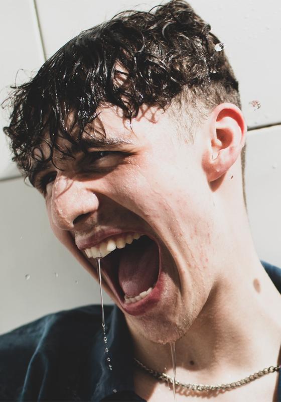 A man with curly hair against a tiled background. His hair is wet and water is dripping from his face.